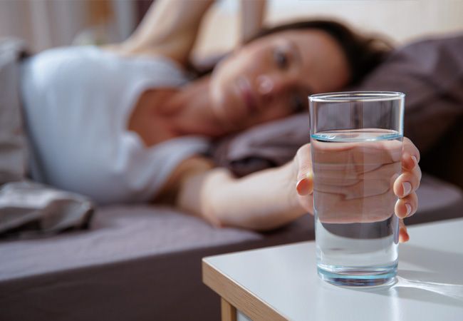 Женщина в белой ночной рубашке лежит в постели, протягивая руку к стакану с водой на прикроватной тумбочке